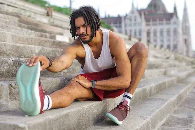 A man in a white tank top is stretching his legs to warm up on concrete stairs