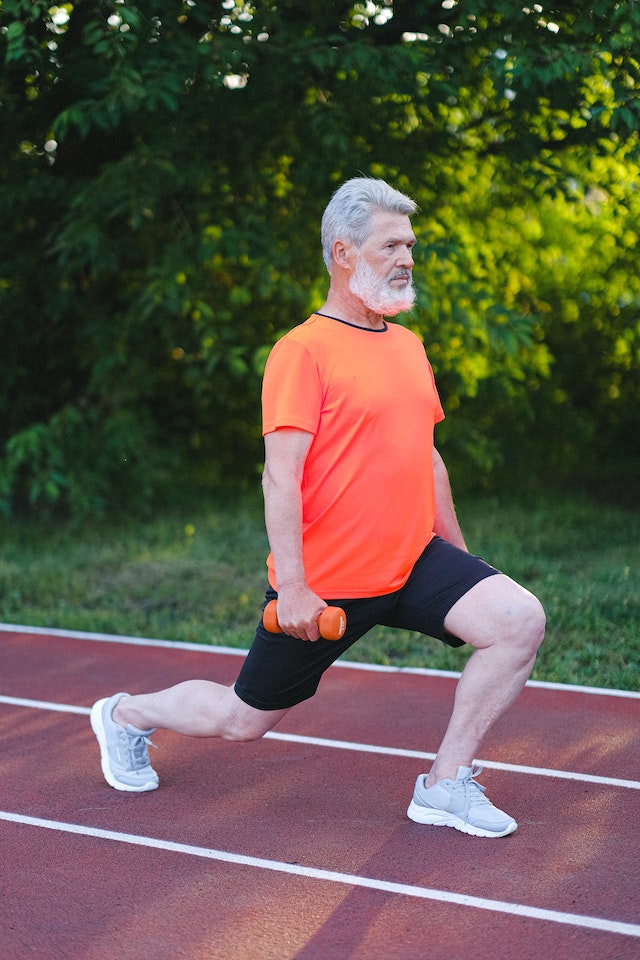 An elderly, fit-looking man in an orange shirt is doing lunges on a red running track, holding one small dumbbell in each hand.