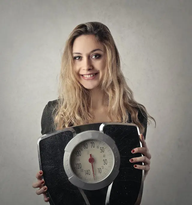 A smiling blonde woman in black shirt holding a black and silver weighing scale.