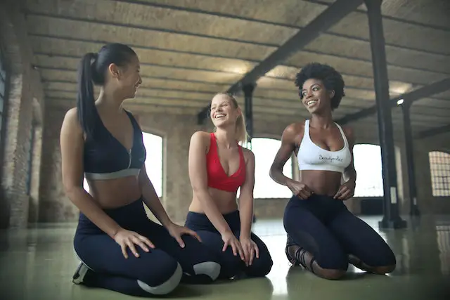 Three beautiful women kneeling on the gym floor and smiling at each other in their fitness attire.