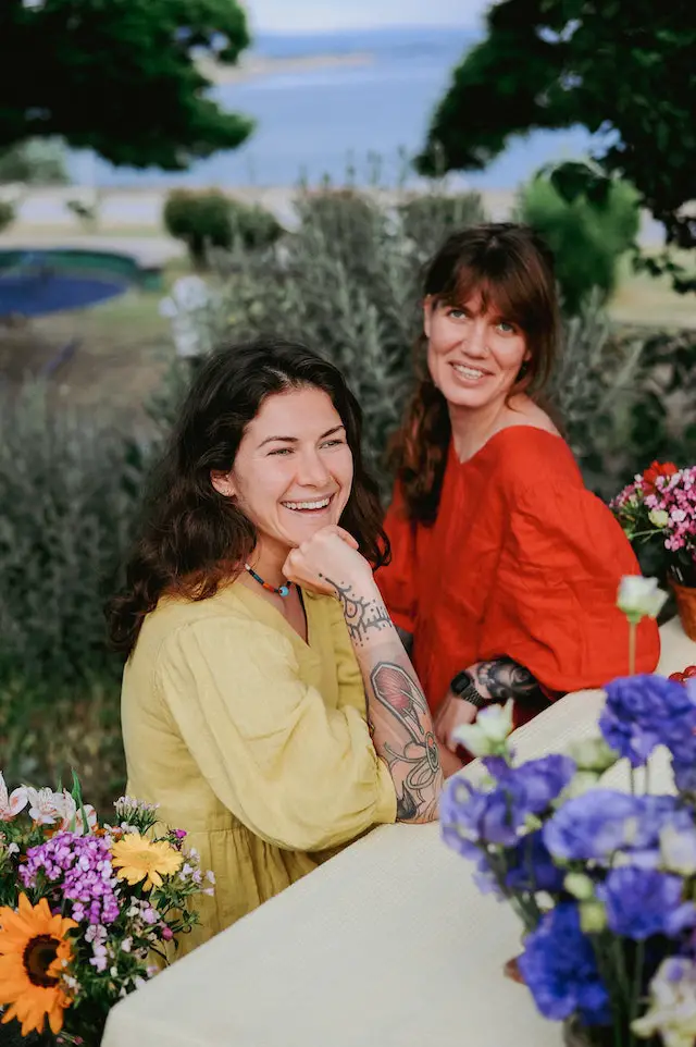 Two smiling women in yellow and red blouse sitting at the table in the garden.