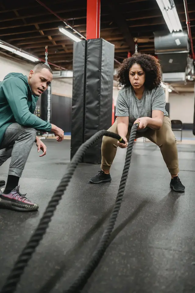 Woman exercising with battle ropes with a trainer watching her.