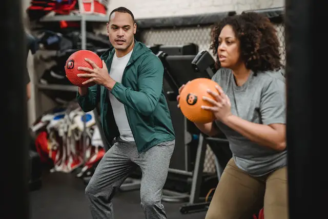 A coach in a green jacket teaches a curly-haired woman how to do a squat workout with an exercise ball.
