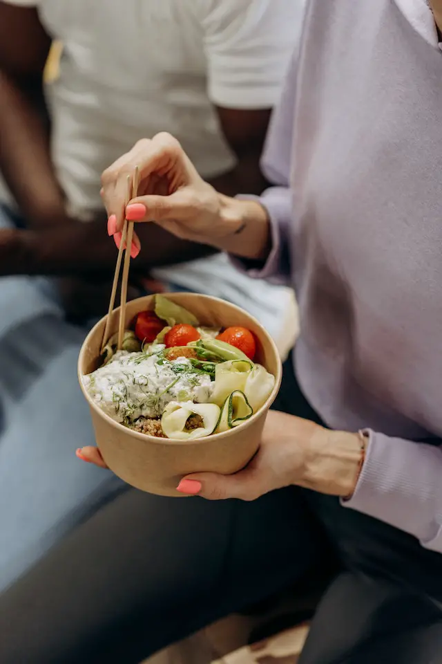 Close-up of a woman's hands holding a bowl of healthy food and using chopsticks to eat from it.