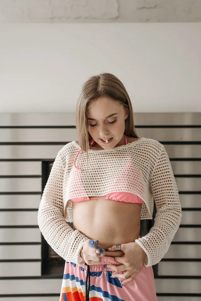 A woman in a white mesh top and pink pants injecting a semaglutide pen on her belly area.