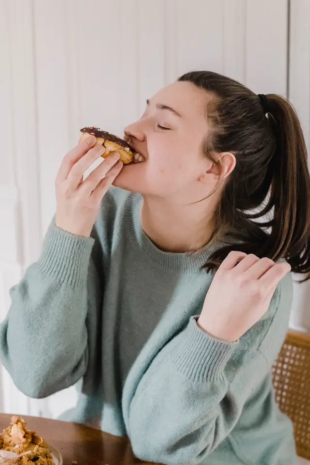 A woman in green shirt, happily eating a chocolate coated donut.