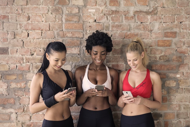 Three women in sports attire holding and reading some information on their smartphones.