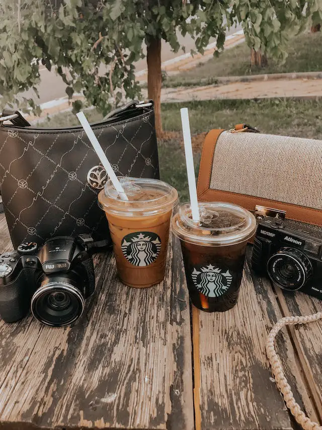 Two Starbucks iced coffees on a wooden table with two bags and two cameras on the side.