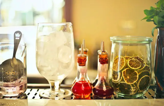 A glass of ice cubes, dried sliced lemons in a glass jar, and a couple of bottles of red syrup on a counter