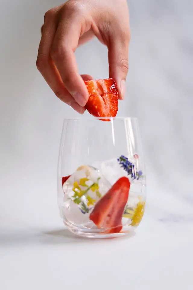 A person adding ice cubes and sliced strawberry in a  clear glass.