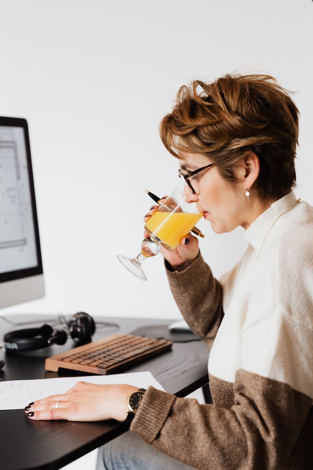 A woman drinking her orange juice in front of her computer.