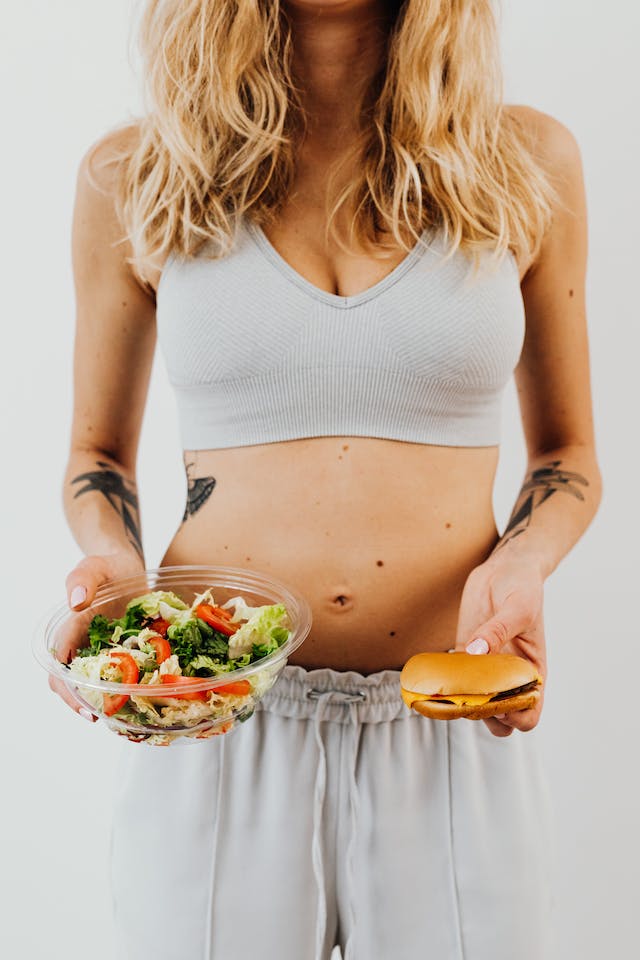 A woman holding a burger and a bowl of salad.