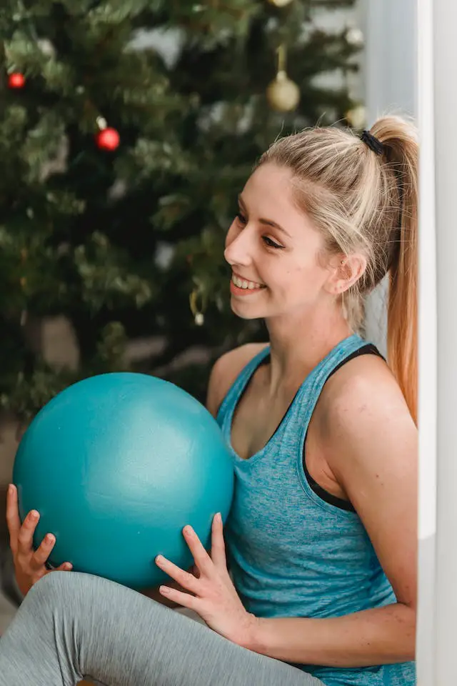 A woman in sports wear sitting beside the Christmas tree while holding a gym ball.