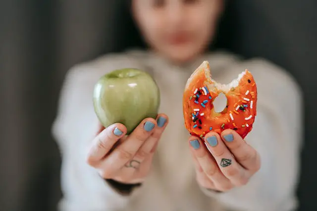 A woman holding out an apple and a bitten sugary donut in front of the camera.