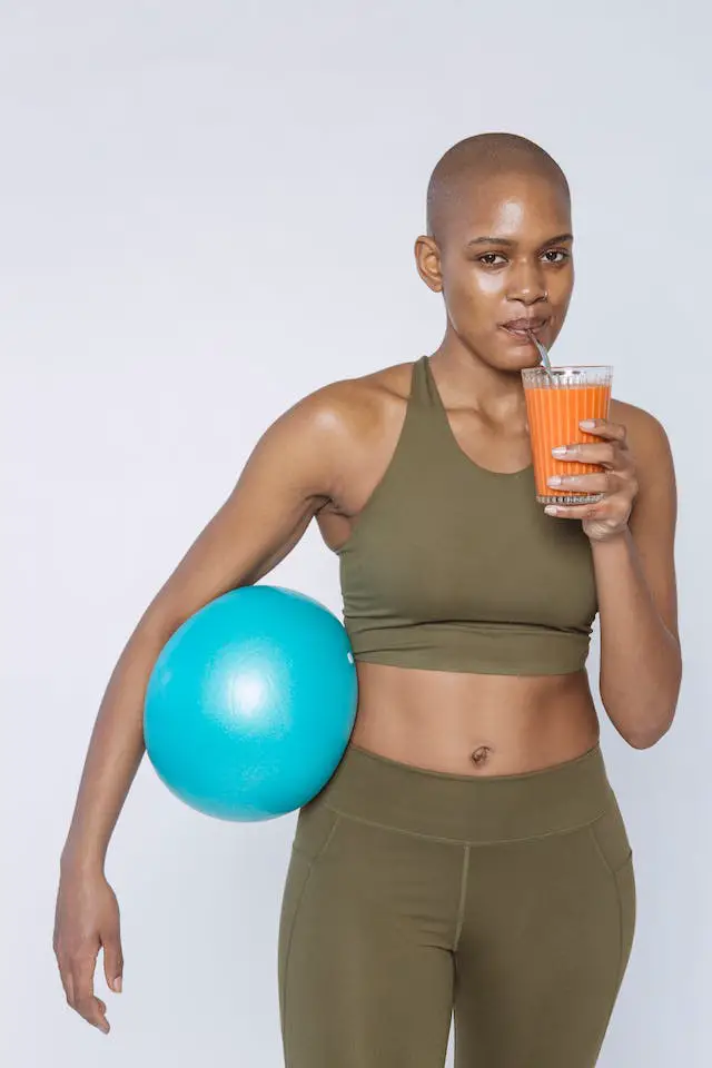 A woman in moss-green sports attire drinking her pink smoothie while holding a gym ball.