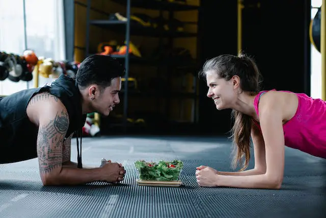 A couple doing planks together face-to-face, smiling at each other with a bowl of salad in between.