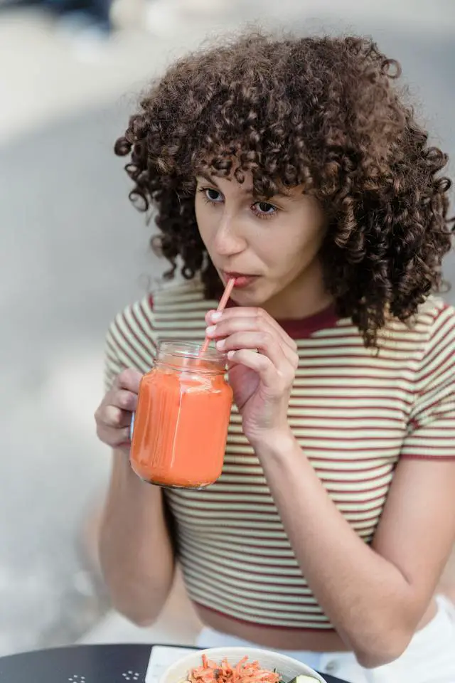 A curly-haired woman drinking a pink smoothie at a street cafe.