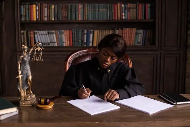A close up shot of a professionally dressed woman writing on her desk with a gavel and a gold balance scale on the side.