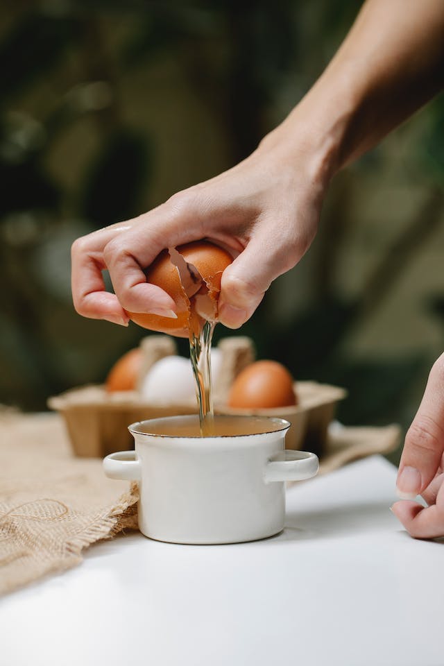 A woman breaking a chicken egg into a small white sauce pan with handle.