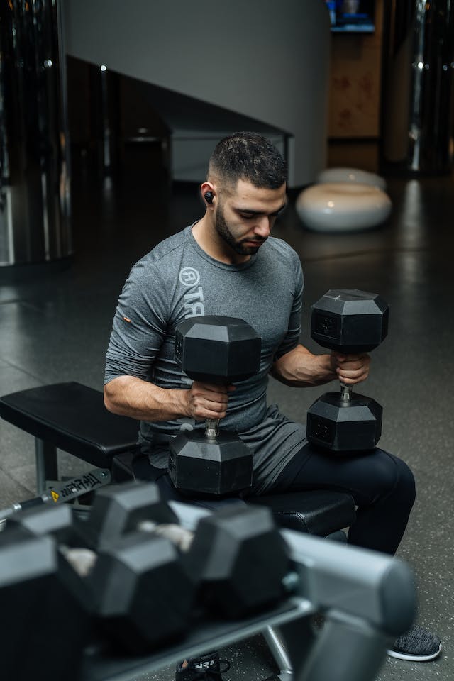 A man in a gray shirt sitting on the bench press with 2 heavy dumbbells standing on his lap.