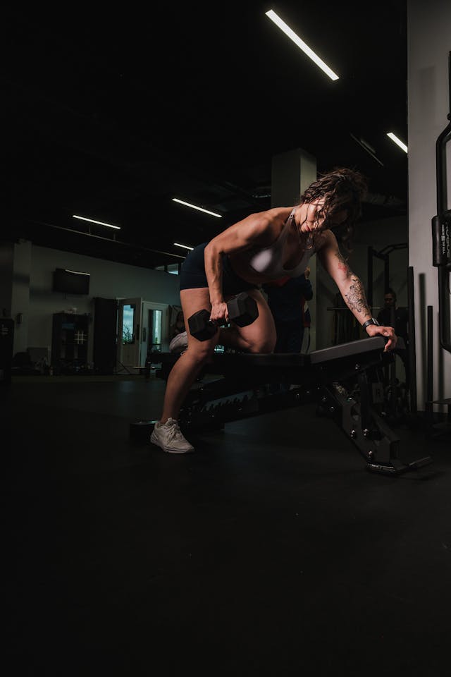 A woman with one knee settled on the gym bench while exercising using a dumbbell.