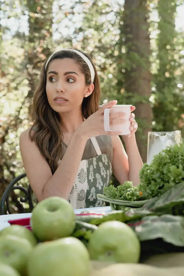 A women holding a white cup while sitting in front of a bunch of green apples and leafy greens on the table.
