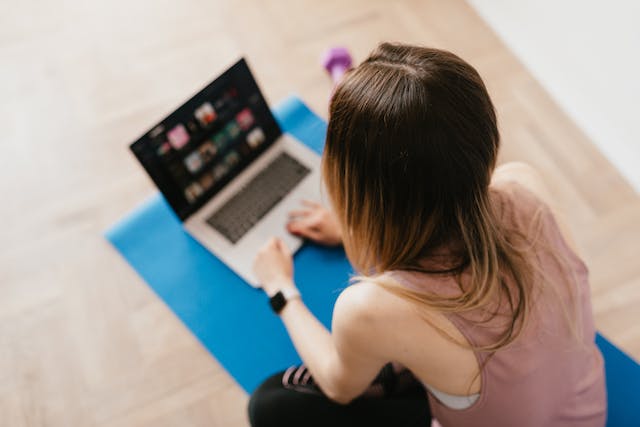 A woman in sports attire sitting on a yoga mat while using her laptop.