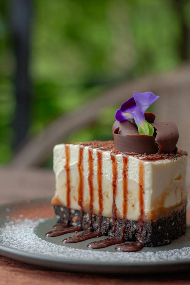 A slice of cheesecake with chocolate icing served on a dark-green plate.