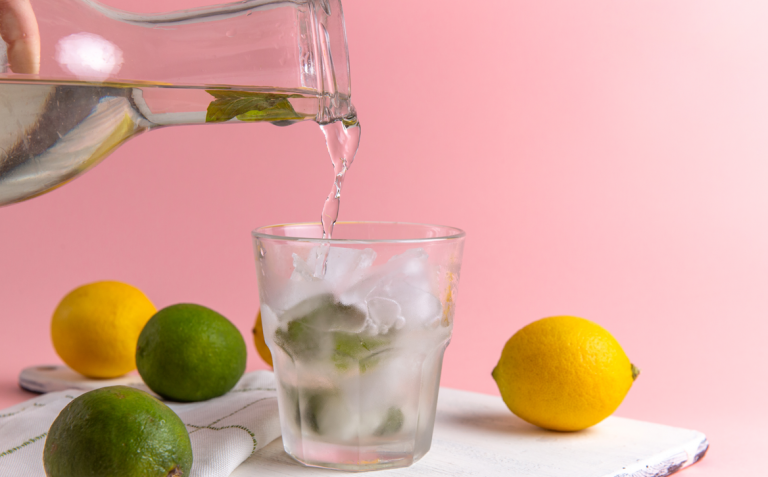 A glass of ice water with mint being poured from a pitcher, surrounded by lemons and limes.