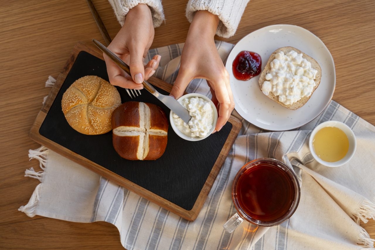 A person spreading cottage cheese on bread rolls with tea, honey, and jam on a wooden table.