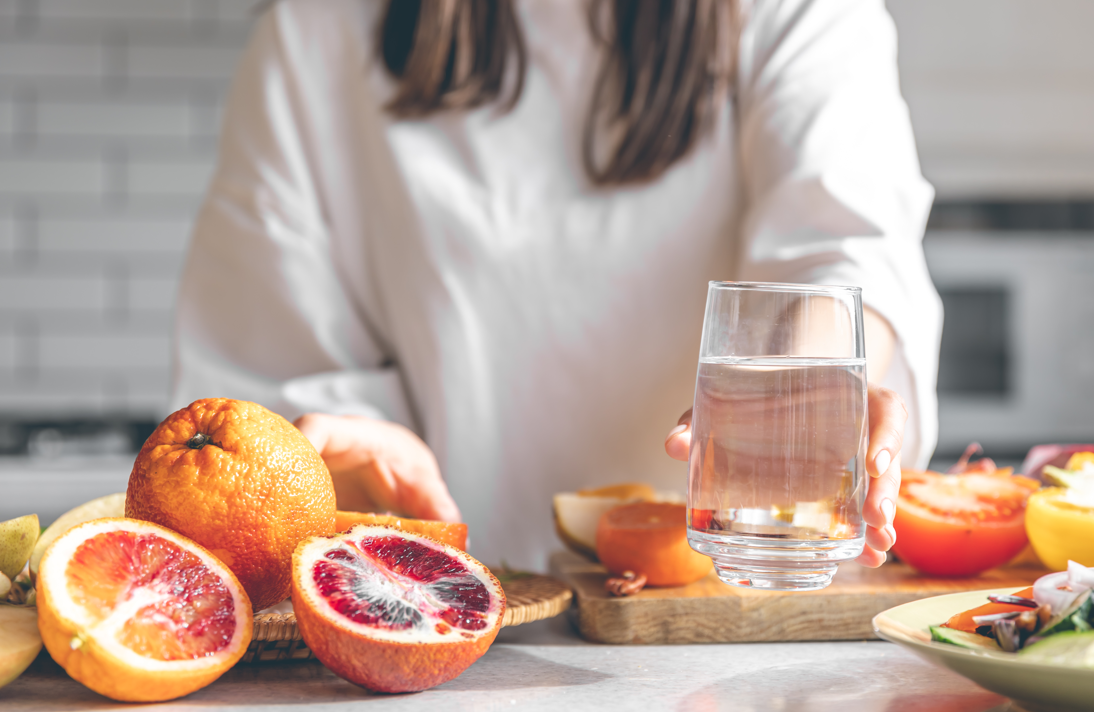 A person holding a glass of water beside a table filled with sliced oranges and other fresh fruit in a kitchen.